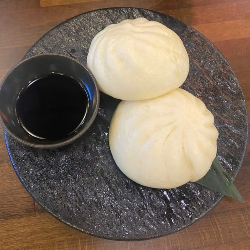 Two fluffy white steamed buns and a small bowl of dark soy sauce on a textured black plate.